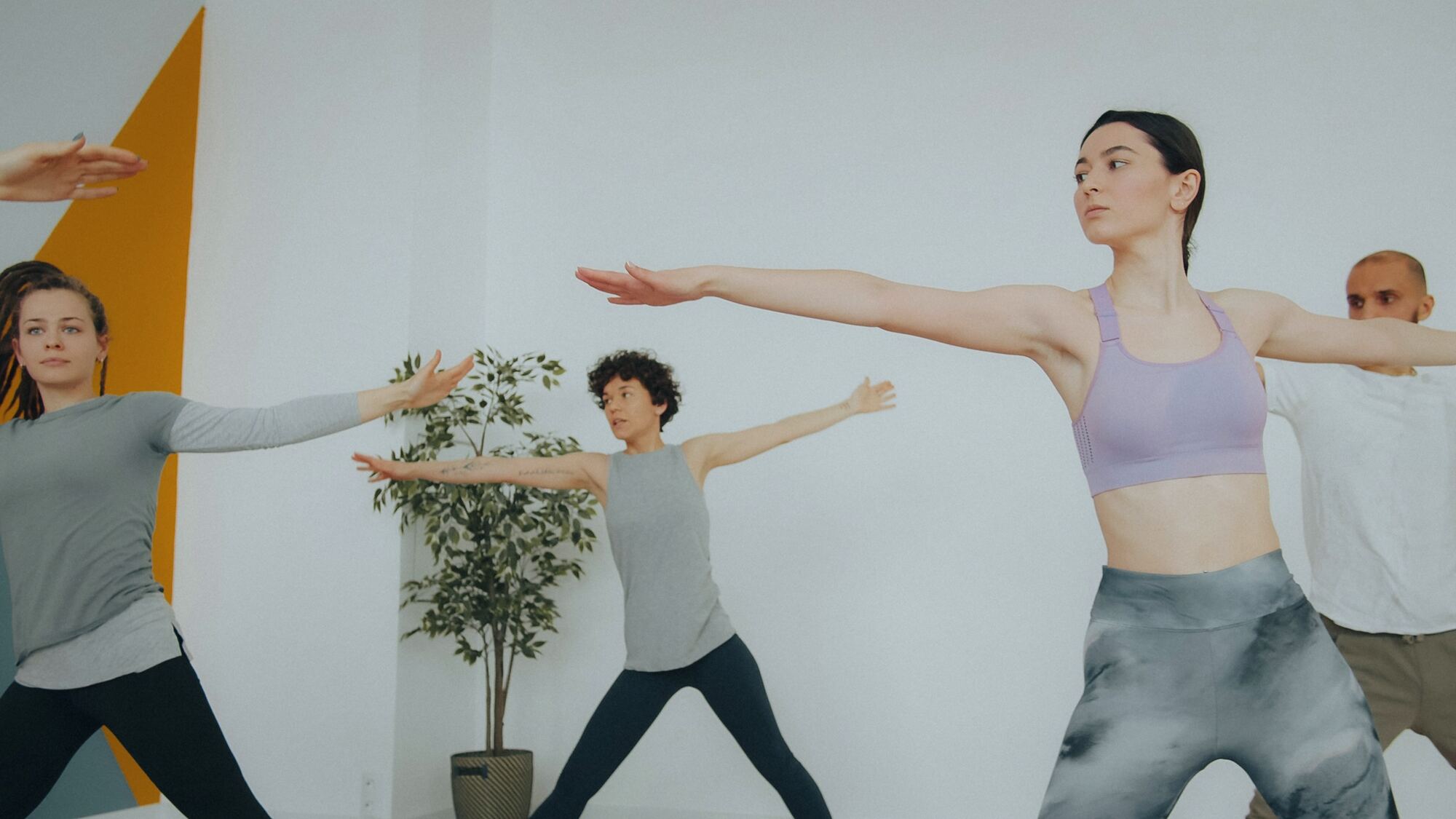 Four people in athletic wear holding a wide-armed standing pose in a bright studio with a plant and a yellow wall