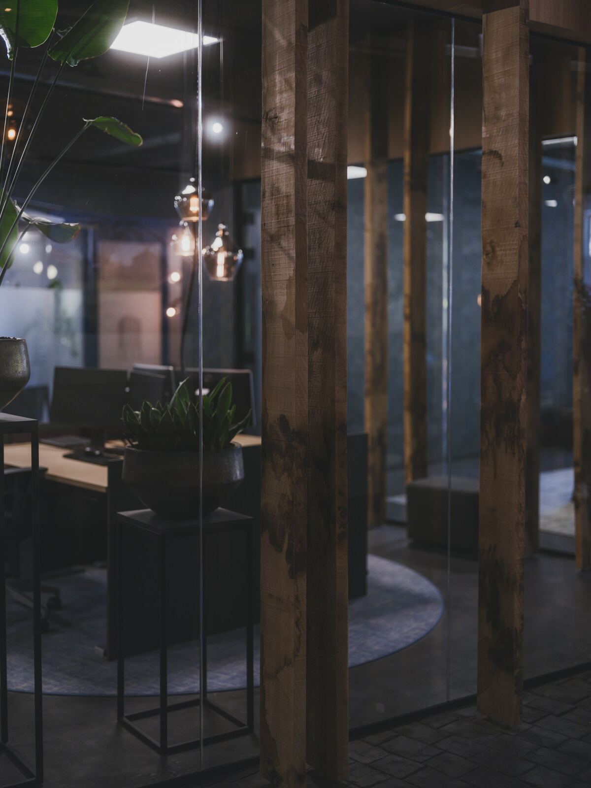 A dimly lit modern office interior seen through a glass partition framed by thick wooden posts, with plants, a desk and pendant lights inside
