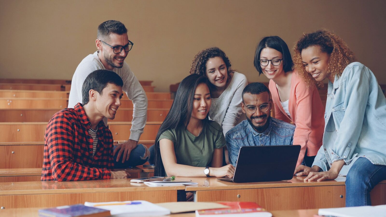 A group of six smiling young adults gather around a laptop in a tiered classroom setting