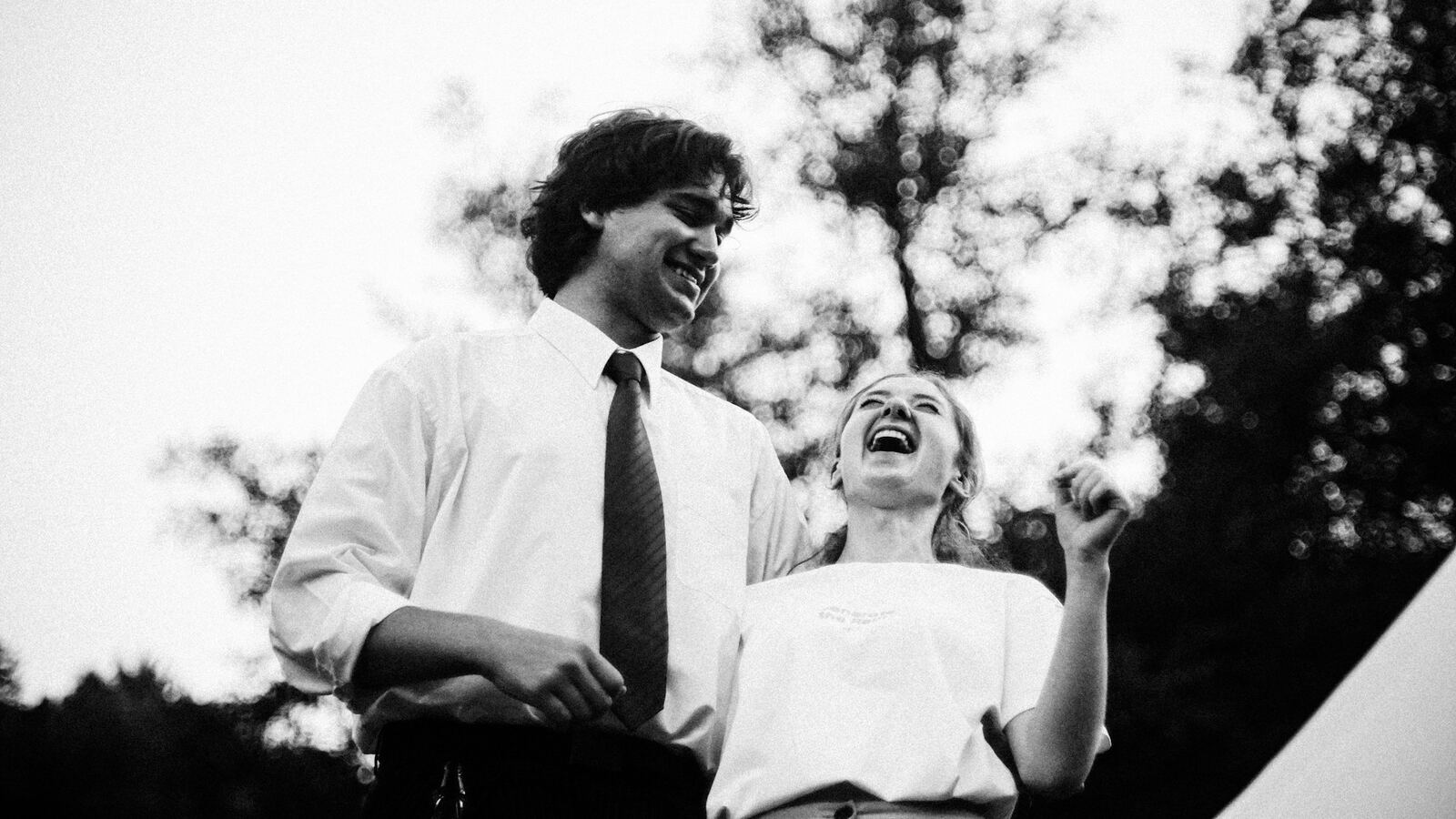A man in a shirt and tie and a woman in a white top laughing together outdoors against a backdrop of trees